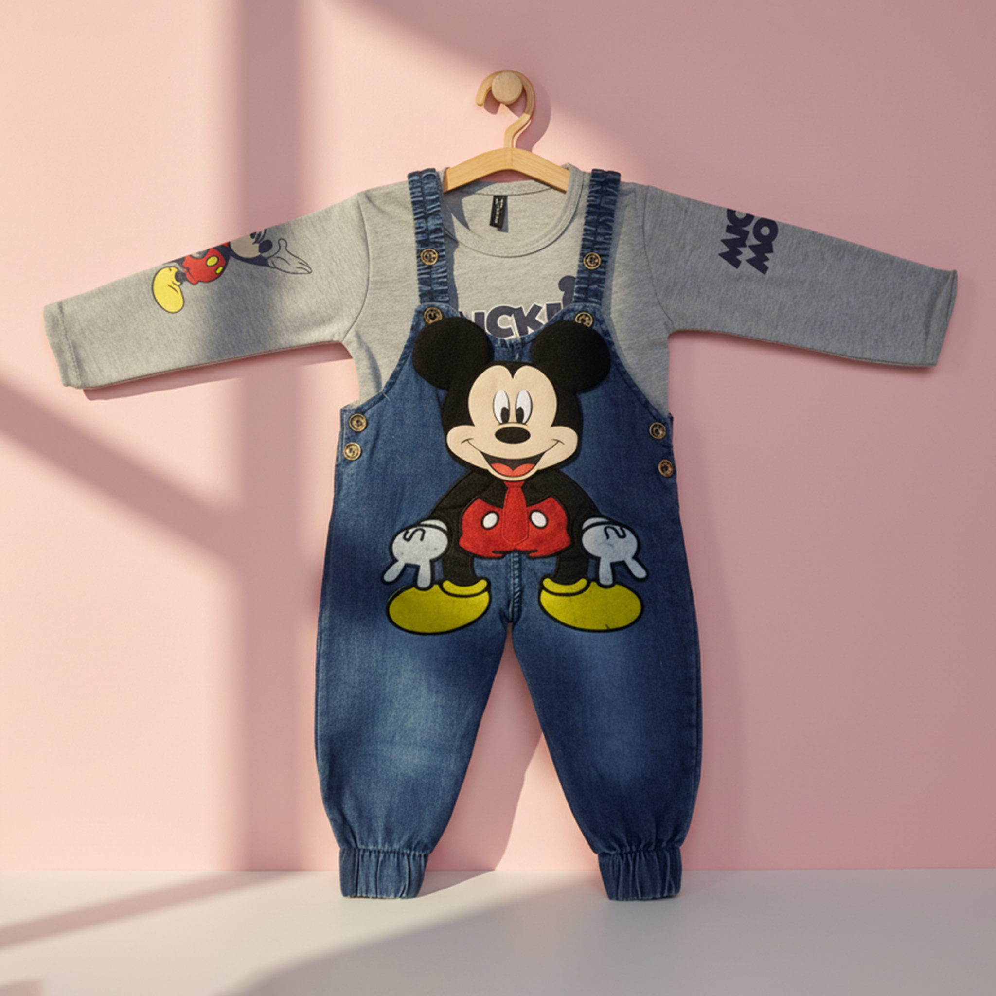 Two children wearing matching Mickey Mouse-themed overalls in a room with a white wall and a shelf with books and plants.