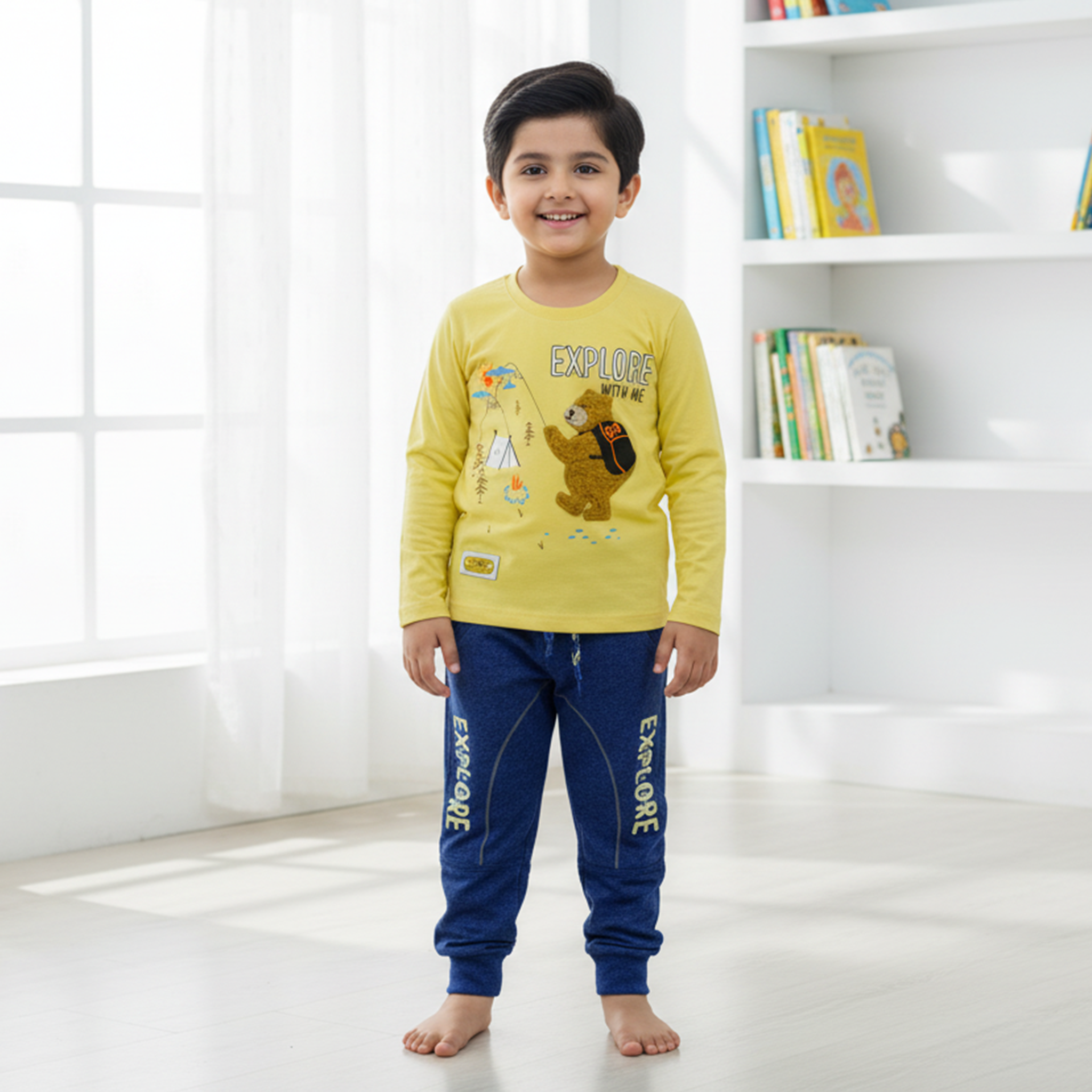 Child wearing a yellow long-sleeve shirt and blue pants with 'EXPLORE' text, standing in a bright room with a bookshelf.